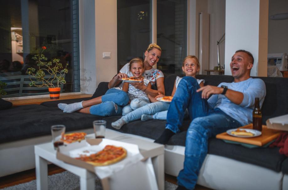 Getty 1271997521 -Caucasian Family Sitting on a Sofa Eating Pizza and Watching TV